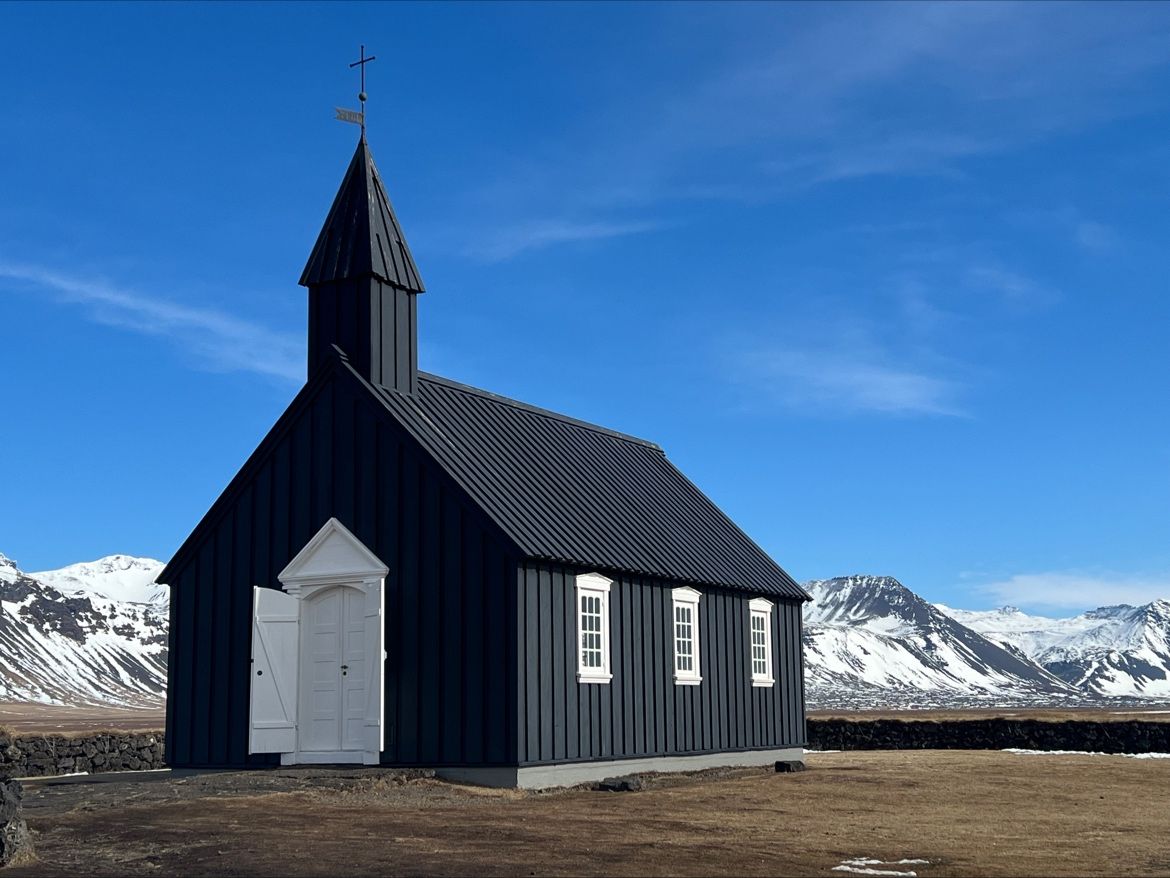 A black wooden church with a white door under a bright blue sky, with snowy mountains behind it, in Iceland.