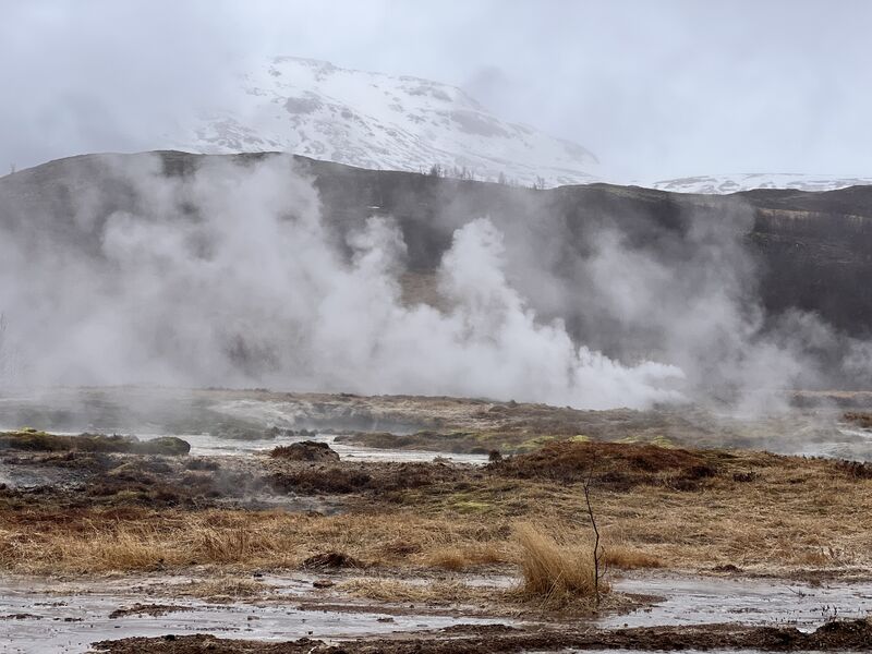 Steam rising from a geothermal field, with a snowy mountain behind.