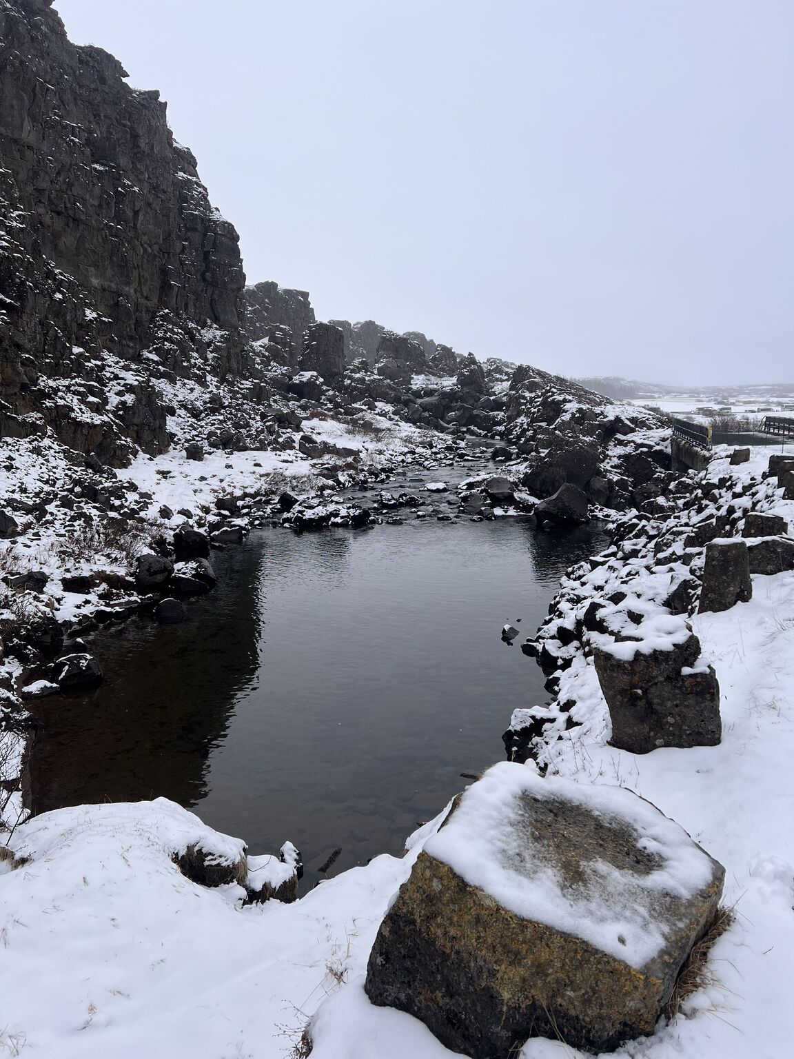 A dark pool of water among snow-covered rocks in a rocky gorge.